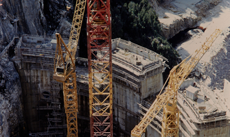 Construction of Gordon Dam, with concrete rising in the shape of the curved dam wall, and red and yellow cranes in the foreground