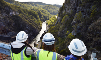 Three young girls looking out downstream of a dam wall, towards a lake winding through two tree-lined shores. The girls are wearing hard hats and high-vis vests.