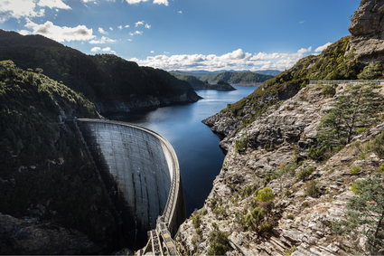 Gordon dam, a curved dam shown in front of an upstream river, a deep blue shine. Rock face lines the right hand side of the river, with trees lining the left hand side. Blue skies above with clouds