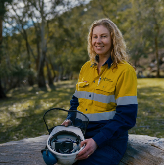 Leah is wearing a high-vis shirt, and sitting on a log facing the camera. She is holding a safety helmet. In the background is grass and trees.