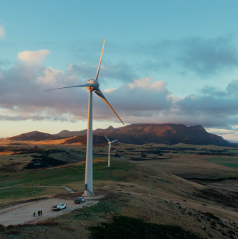 A wind turbine standing amongst green fields, with the sun setting in the background. Blue dusty skies and clouds surround the top of the turbine. A smaller wind turbine can be seen in the background also.